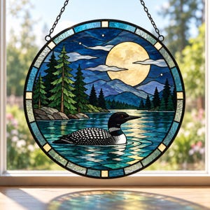 May include: A circular stained glass suncatcher featuring a loon swimming on a lake at night. The scene includes a full moon, silhouetted pine trees, and blue mountains. The glass is framed in a black metal border with a hanging chain attached.