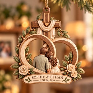 May include: A hanging ornament featuring a wooden cross with a draped cloth and a small heart, framing a couple from behind. Two interlocking rings surround the figures, accented by green leaves and white roses. Text reads: SOPHIE & ETHAN JUNE 18, 2016.