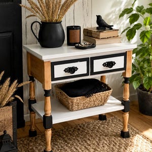 May include: A wooden side table with a white top, two drawers featuring black frames and ornate black knobs, and a lower shelf. The table has four turned legs with black accents and natural wood tones. A woven basket sits on the lower shelf.