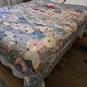 May include: A patchwork quilt featuring a variety of fabric squares in shades of blue, tan, white, and red. The squares are joined with small, knotted fabric ties at each corner, creating a textured surface across the entire bed covering.
