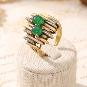 Könnte beinhalten: Ein goldfarbener Ring mit einem strukturellen Design aus mehreren parallelen Stäben, zwei ovalen grünen Cabochon-Steinen und kleinen klaren Pavé-gefassten Kristallen, die entlang der Metalloberflächen eingelassen sind, auf einem zylindrischen Ständer.