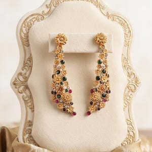 Puede incluir: Un par de pendientes colgantes de tono dorado con un diseño curvo inspirado en flores. Los pendientes están adornados con pequeñas piedras preciosas multicolores en tonos rojos, verdes y transparentes, engastadas en metal sobre un expositor crema.