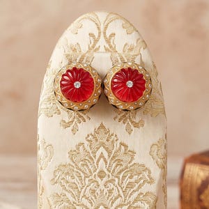 Puede incluir: Un expositor de joyas de forma ovalada cubierto de tela color crema con un patrón de damasco dorado. El soporte presenta dos pendientes redondos de color rojo con forma de flor, piedras claras centrales y una base de metal bronce.