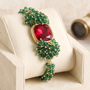 Può includere: Un bracciale caratterizzato da una grande pietra preziosa rossa quadrata e sfaccettata incastonata in una cornice dorata, affiancata da grappoli di piccole perline rotonde verde scuro. Le perline sono disposte in gruppi densi e strutturati lungo la catena dorata.