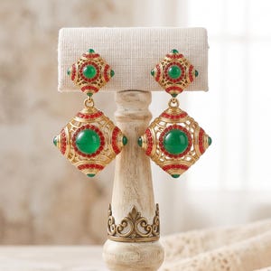 Puede incluir: Un par de pendientes colgantes dorados con un diseño en forma de diamante, una piedra cabujón verde central, rodeada de pequeños cristales rojos y filigrana metálica, expuestos en un soporte para pendientes de tela color crema.