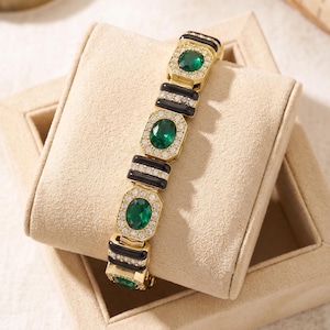 May include: A gold-toned link bracelet featuring alternating oval-cut emerald green stones and rectangular black enamel segments. Each green stone is surrounded by a halo of small clear crystals, and the black segments are accented with clear crystals.