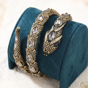 May include: A coiled snake bracelet in an antique gold tone finish featuring a textured scale pattern. The design is embellished with multiple teardrop-shaped clear crystals along the body and head, with small round crystals accenting the head.