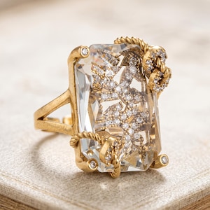 May include: A gold-toned ring featuring a large, rectangular, clear faceted stone. The stone contains an intricate, embedded metallic branch-like design encrusted with tiny white crystals. The gold band has a twisted rope texture.
