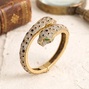 Può includere: Un bracciale rigido incernierato color oro a forma di pantera con la testa appoggiata sul cerchio. La superficie è incastonata con piccoli cristalli trasparenti e macchie geometriche nere, con occhi di cristallo verde brillante sulla testa.