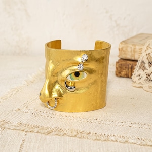 Op de afbeelding: Een goudkleurige manchetarmband met een gebeeldhouwde menselijke neus en één oog met een groene iris. Het ontwerp bevat kleine heldere kristallen accenten boven het oog, een zilverkleurige traanvorm onder het oog en een kleine met kristallen bezette neusring.