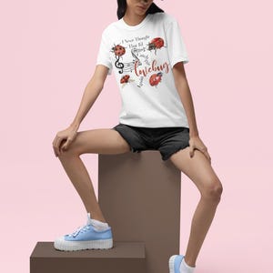 Pode incluir: Uma t-shirt branca de manga curta com um gráfico de joaninhas vermelhas, notas musicais e o texto "I Never Thought That I'd Catch This Lovebug Again". A peça é combinada com um boné preto, calções de ganga pretos e ténis azul-claros.