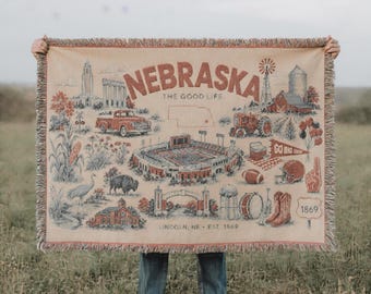 Puede incluir: Una manta tejida con fondo crema e ilustraciones en color óxido de monumentos de Nebraska, un estadio de fútbol, un tractor, una camioneta, un bisonte, botas de vaquero y el texto Nebraska The Good Life Lincoln NE EST 1869.