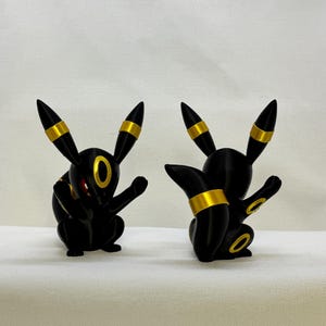 May include: Two black and gold figurines of a four-legged creature with long pointed ears. The figures feature yellow ring markings on the ears, forehead, tail, and limbs. One figure faces forward showing red eyes, while the other is turned away.