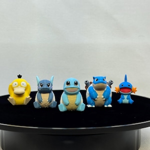 May include: A set of five small, colorful plastic character figurines arranged in a row. The figures include a yellow duck-like creature, a blue creature with pointed ears, a light blue turtle, a larger blue turtle with cannons, and a small blue creature with orange fins.