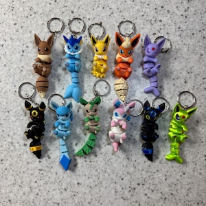 May include: A collection of eleven articulated keychains featuring various fictional creatures in distinct colors including brown, blue, yellow, orange, purple, black, green, and pink. Each figure has a segmented tail and a metal ring for attachment.