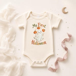May include: A cream-colored short-sleeve baby bodysuit featuring a graphic of a white ghost juggling three orange pumpkins. The text "My First Halloween" is printed in brown and green lettering above and below the ghost illustration.