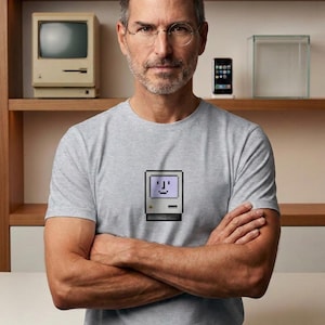 May include: A light gray short-sleeved t-shirt featuring a centered pixel art graphic of a vintage beige computer with a smiling face on the screen. The design includes a small multicolored square logo at the bottom left of the computer illustration.