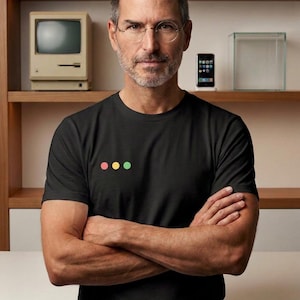 May include: A black short-sleeved t-shirt featuring three small, horizontally aligned circles in light red, yellow, and light green on the left chest. The shirt has a crew neckline and is worn by a person with short hair and round glasses.