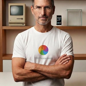 Puede incluir: Una camiseta blanca de manga corta con un gráfico circular colorido de diseño en espiral y el texto Still Processing impreso en gris debajo. La camiseta la lleva un hombre de pelo corto y gafas redondas, de pie con los brazos cruzados.