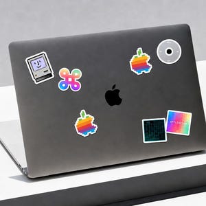 Może przedstawiać: Ciemnoszara pokrywa laptopa z czarnym logo Apple pośrodku i sześcioma naklejkami: spikselizowany komputer, kolorowy symbol polecenia, dwa spikselizowane tęczowe jabłka, okrągłe kółko nawigacyjne, płytka drukowana i kwadrat z gradientem.