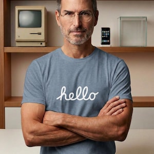 Könnte beinhalten: Ein blaues, meliertes Kurzarm-T-Shirt mit dem Wort hello in weißer Schreibschrift auf der Brust. Das Shirt hat einen Rundhalsausschnitt und eine normale Passform und besticht durch ein schlichtes Design mit einer klaren, minimalistischen Optik.