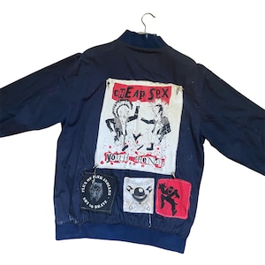 Puede incluir: Una chaqueta bomber azul oscuro con un gran parche rectangular en la espalda con el texto CHEAP SEX y YOUTH OF ENDS, que muestra dos figuras con crestas. Debajo hay tres parches más pequeños: uno negro circular, uno blanco y uno rojo.