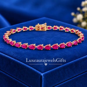 Könnte beinhalten: Ein goldfarbenes Armband mit einer durchgehenden Reihe birnenförmiger, leuchtend pinker Edelsteine in Metallfassungen. Das Schmuckstück verfügt über einen sicheren Kastenschloss-Verschluss. Der Text LuxeaurajewelsGifts ist unter dem Armband sichtbar.
