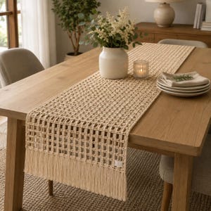 Pode incluir: Um caminho de mesa de macramé bege com um padrão de grelha aberta e franjas longas nas extremidades. O caminho está estendido sobre uma mesa de jantar de madeira, exibindo a construção intrincada de cordão atado e o design tecido.