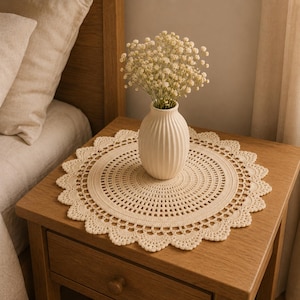 May include: A round, cream-colored crocheted doily featuring a detailed, intricate openwork pattern with a scalloped edge. The doily is topped with a white, ribbed ceramic vase filled with a bouquet of small, delicate white baby's breath flowers.