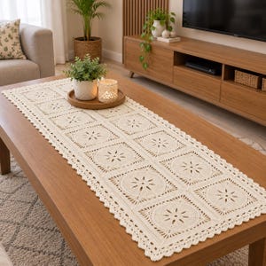Pode incluir: Um caminho de mesa retangular de crochê na cor creme, apresentando um padrão repetitivo de motivos quadrados com centros florais circulares e bordas festonadas. O caminho está estendido sobre uma superfície de madeira, exibindo textura.
