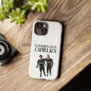 May include: A white smartphone case featuring the black text "LOS FABULOSOS CADILLACS" above a graphic of two individuals wearing black racing-style jumpsuits with white accents. The case has a cutout for a dual-lens camera system on the back.