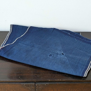 May include: A folded piece of dark blue indigo-dyed fabric with a visible woven texture. The textile features a distinct selvedge edge with thin white and red stripes, and two small holes are present in the centre of the folded material.