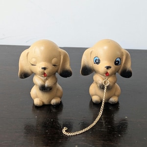 以下が含まれることがあります： 耳と足先が黒い、日焼けした色の陶器製の犬の置物2体。1体はまつ毛が描かれた閉じられた目をしており、もう1体は開いた青い目をしています。開いた目の犬の首には、細い金色の金属製のチェーンが繋がっています。