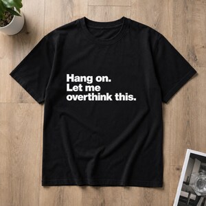 May include: A black short-sleeved t-shirt featuring white text printed on the chest that reads, Hang on. Let me overthink this. The garment has a crew neckline and a standard fit, presented as a flat lay item with a simple design.