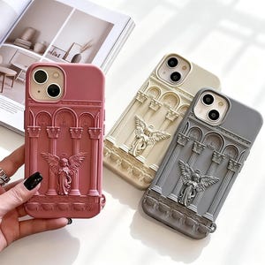 May include: Three smartphone cases in dusty rose, cream, and slate gray. Each case features a raised, three-dimensional relief design depicting a central winged angel figure standing between classical architectural columns and arched structures.