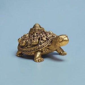 May include: A gold-coloured metal turtle figurine featuring intricate relief carvings of deities on its shell, topped with a small pyramid-shaped structure. The turtle has a textured skin surface, four legs, and a head extended forward.