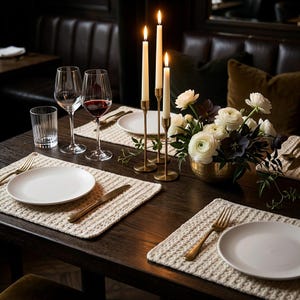 Puede incluir: Una mesa de comedor de madera oscura con manteles individuales texturizados de color crema, platos de cerámica blanca, cubiertos dorados, cristalería transparente, un candelabro de latón con tres velas blancas y un cuenco de latón con ranúnculos.