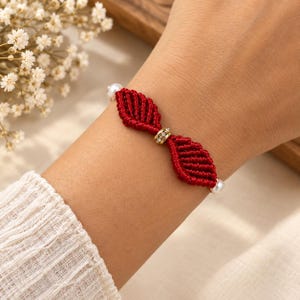 Könnte beinhalten: Ein Makramee-Armband mit zwei dunkelroten, geknoteten blattförmigen Elementen, die durch eine goldfarbene Perle mit kleinen klaren Kristallen verbunden sind. Das Armband ist an beiden Enden mit einer weißen Perle versehen.