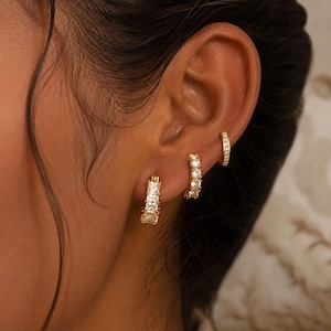 May include: Three gold hoop earrings worn in a single ear. The largest hoop features a row of rectangular baguette-cut clear stones. The middle hoop is set with a row of round clear stones. The smallest hoop is a thin, plain gold band.