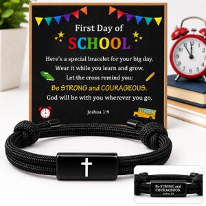 May include: A black cord bracelet featuring a rectangular metal charm with a white cross on the front and the text "Be STRONG and COURAGEOUS. Joshua 1:9" on the back, displayed with a card titled "First Day of SCHOOL" with colorful bunting.