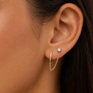 Op de afbeelding: Een gouden kettingoorbel bestaande uit een kleine ring met ingebedde heldere stenen, verbonden door een fijne gouden ketting met een enkele oorsteker met een heldere steen. Het sieraad wordt gedragen in twee gaatjes in de oorlel.
