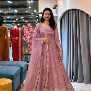 Puede incluir: Un vestido Anarkali malva hasta el suelo con mangas largas, cuello redondo y bordados dorados intrincados en el dobladillo y los puños. El vestido incluye una dupatta transparente a juego con un borde dorado decorativo y motivos florales.