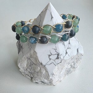 May include: Two bracelets featuring round beads in shades of blue, dark gray, and light green, woven with metallic wire. One bracelet uses gold-toned wire, while the other uses silver-toned wire, both displaying a repeating crisscross pattern.