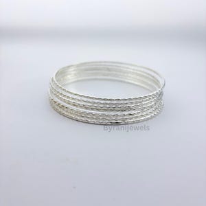 May include: A set of five stacked silver bangles, each featuring a delicate, repeating scalloped or wavy texture along the outer surface. The bracelets are metallic, reflective, and circular, with the text Byranijewels visible in the lower right.