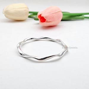 Może przedstawiać: Polerowana srebrna bransoletka typu bangle o ciągłym, falistym wzorze. Okrągła bransoletka ma gładką, odblaskową metaliczną powierzchnię. Napis Byrenijewels jest widoczny małą, jasną czcionką po prawej stronie bransoletki.