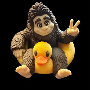 May include: A 3D printed figurine of a dark brown, furry creature with a light-colored face, hands, and feet. The creature is sitting on a yellow rubber duck, making a peace sign with one hand and smiling with a friendly, cartoon-like expression.