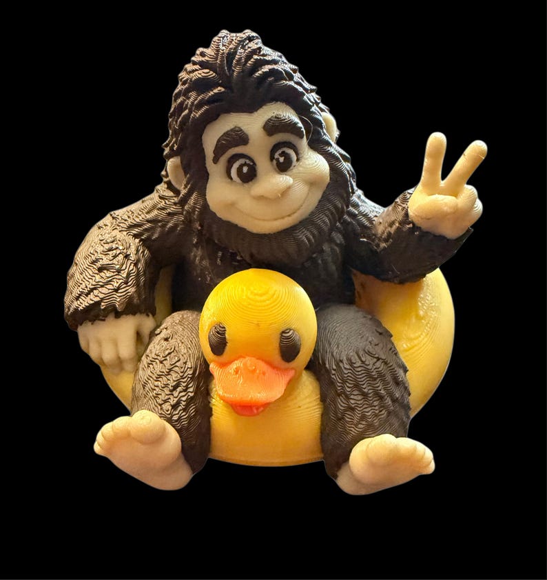 May include: A 3D printed figurine of a dark brown, furry creature with a light-coloured face, hands, and feet. The creature is sitting on a yellow rubber duck, making a peace sign with one hand and smiling with a friendly, cartoon-like expression.