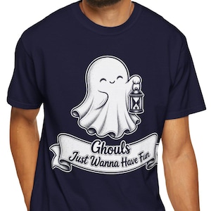 May include: A navy blue short-sleeve t-shirt featuring a white graphic of a smiling ghost holding a lantern. Below the ghost is a white banner with the black text "Ghouls Just Wanna Have Fun" written in a stylized, cursive script font.