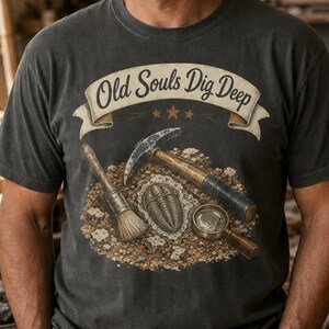 Op de afbeelding: Een antracietgrijs T-shirt met korte mouwen en een grafische print met de tekst Old Souls Dig Deep op een banner. Het ontwerp bevat een geologische hamer, een kwast, een vergrootglas en een trilobiet-fossiel op een textuurrijke achtergrond.