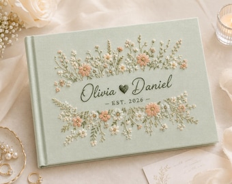 Könnte beinhalten: Ein helles, salbeigrünes, stoffbezogenes Fotoalbum mit aufwendiger Blumenstickerei in Pfirsich-, Weiß- und Grüntönen. In der Mitte sind die Namen Olivia und Daniel, getrennt durch ein Herz, sowie der Text EST. 2026 darunter eingestickt.